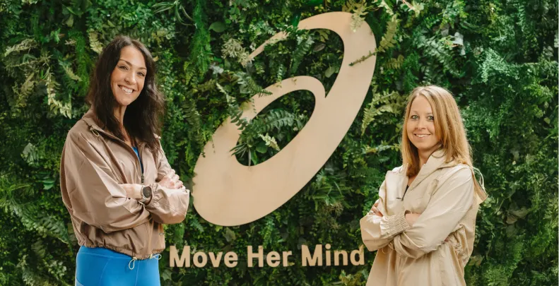 Two women standing in front of a living wall with a large ASICS "A" and "Move Her Mind"